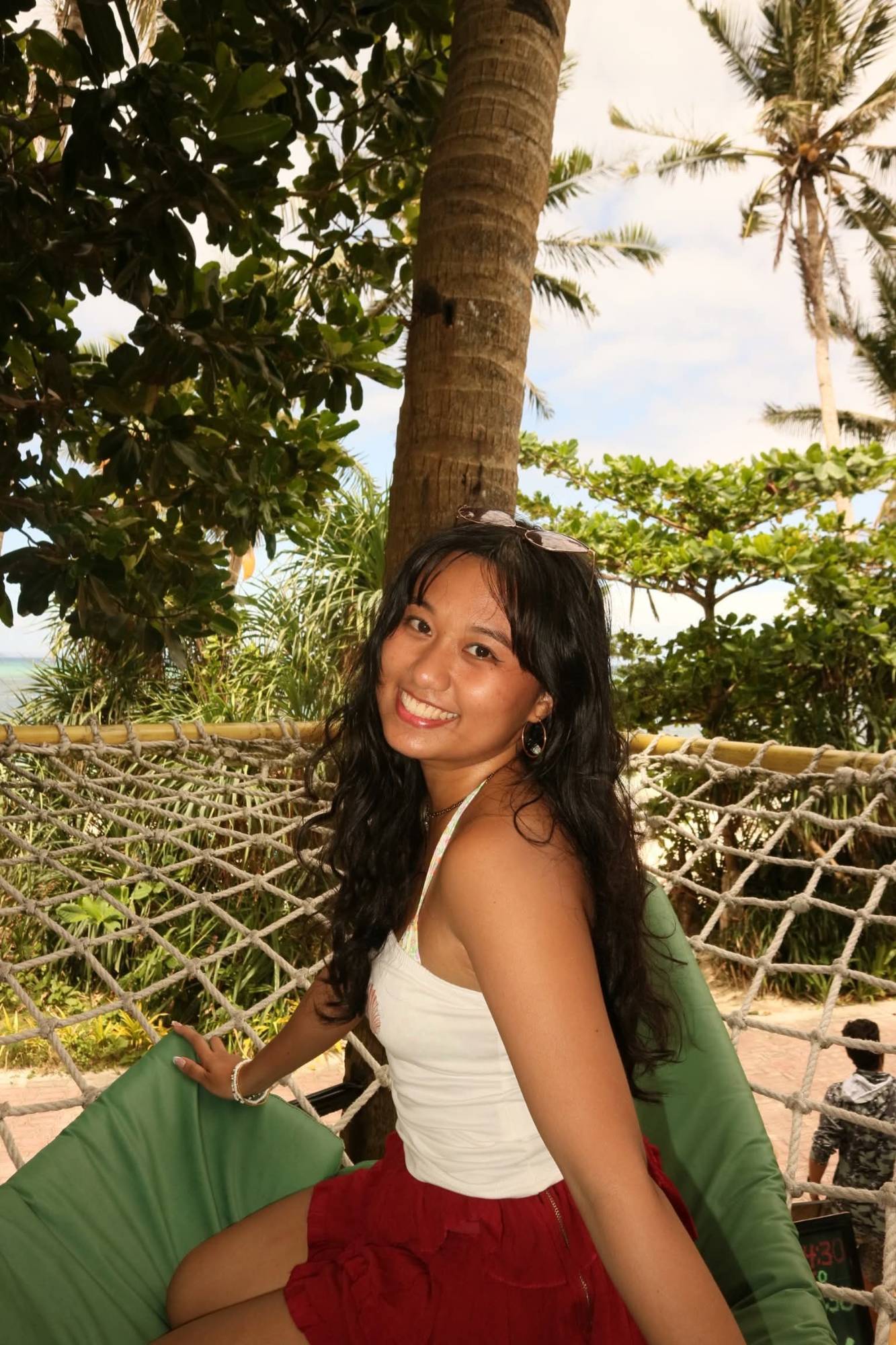 Elle smiling in a rope hammock under palm trees, seen through an airplane window